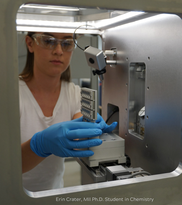 Erin Crater A woman wearing safety glasses is seen through the window of a large laboratory instrument.