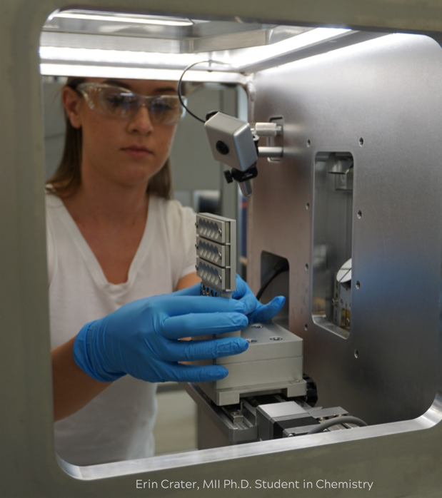 A woman wearing safety glasses is seen through the window of a large laboratory instrument.